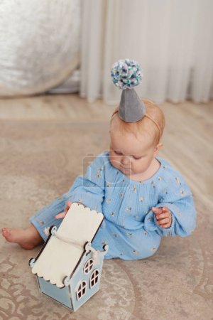 Foto de Adorable baby 1 year old girl dressed in blue floral dress and birthday cone hat with pompon playing and exploring her gift, wooden house by floor with fluffy carpet. Children day. Happy birthday. - Imagen libre de derechos
