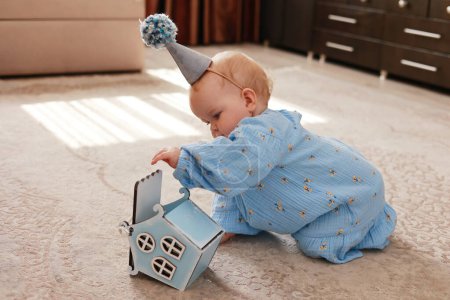 Foto de Adorable baby 1 year old girl dressed in blue floral dress and birthday cone hat with pompon playing and exploring her gift, wooden house by floor with fluffy carpet. Children day. Happy birthday. - Imagen libre de derechos