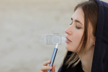 Electronic cigarette technology. Young woman smokes and releases steam from hybrid cigarette device that uses real tobacco refills with a heating pad, tobacco heating system. Bad unhealthy habit.