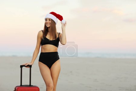 Téléchargez les photos : Slim jeune femme souriante dans le chapeau du Père Noël et bikini noir se tient avec valise rouge sur le bord de la mer. Vacances de Noël et Nouvel An. Célébration des vacances d'hiver à l'étranger sous les tropiques. Voyage de dernière minute. - en image libre de droit