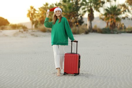 Téléchargez les photos : Joyeux jeune femme dans le chapeau du Père Noël et sweat-shirt vert se tient avec valise rouge sur la plage avec paumes. Vacances de Noël et Nouvel An. Célébrer les vacances d'hiver à l'étranger dans les tropiques.. - en image libre de droit