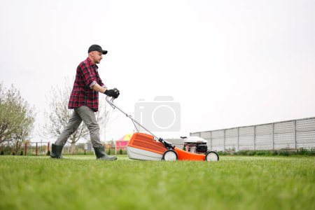 Photo for View of man in casual clothes mows lawn with lawn mower at backyard of his house. Husband takes care of garden on spring cloudy day. Modern gasoline garden equipment. Landscaping work. - Royalty Free Image