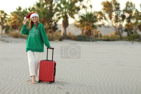 Téléchargez les photos : Joyeux jeune femme dans le chapeau du Père Noël et sweat-shirt vert se tient avec valise rouge sur la plage avec paumes. Vacances de Noël et Nouvel An. Célébrer les vacances d'hiver à l'étranger dans les tropiques.. - en image libre de droit