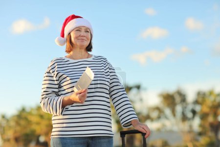 Téléchargez les photos : Femme mûre indépendante dans le chapeau de Noël Père Noël, chemise rayée détient passeport, billet d'embarquement, valise sur le bord de la mer avec des paumes de fond célèbre le Nouvel An vacances dans un complexe exotique. - en image libre de droit