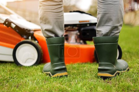 Photo for Cropped photo of male gardener legs in rubber boots stands on cut green grass lawn at backyard of house. Modern electric cordless lawnmower on back. Landscaping industry theme - Royalty Free Image