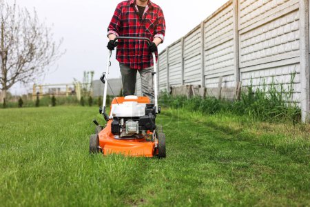 Photo for View of man in casual clothes mows lawn with lawn mower at backyard of his house. Husband takes care of garden on spring cloudy day. Modern gasoline garden equipment. Landscaping work. - Royalty Free Image