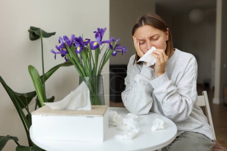 Sick unhealthy allergic young woman covering nose with paper tissue has runny stuffy nose, sneezes from iris flowers pollen at kitchen home. Girl with flu, itching or cough, seasonal allergy, rhinitis