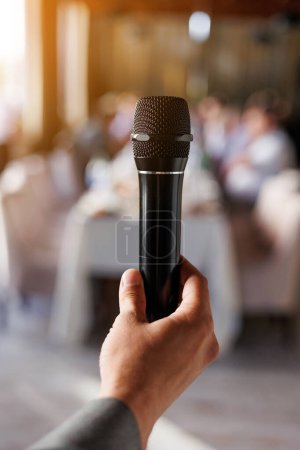 Téléchargez les photos : Une main masculine méconnaissable tient un microphone noir, un maître de cérémonie ou un discours de showman devant le public lors d'un événement. Sur le fond s'asseoir les invités à la table. Fête et événements festifs concept - en image libre de droit