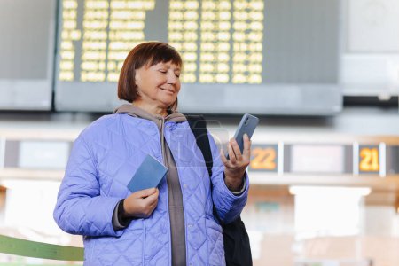 Photo for Middle aged woman traveler with backpack holds passport, uses cellphone at public airport terminal after check in at airline services desks. Active retirees. Travelling, vacation, tourism concept - Royalty Free Image