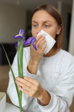 Mujer joven alérgica sostiene flor de iris, cubre la nariz con papel tejido tiene secreción nasal, estornudos de flores polen en la cocina casa por mesa. Niña con gripe, picor o tos, alergia estacional, rinitis.