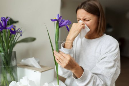 Allergic young woman holds iris flower, covers nose with paper tissue has runny nose, sneezes from flowers pollen at kitchen home by table. Girl with flu, itching or cough, seasonal allergy, rhinitis.