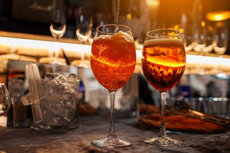 Foto de Dos vasos de cóctel Aperol spritz en el mostrador del bar. Elaboración de bebidas típicas italianas alcohólicas, aperitivo elaborado con vino blanco espumoso Prosecco, hielo y rodajas de naranja fresca. Mezcla de líquido naranja - Imagen libre de derechos