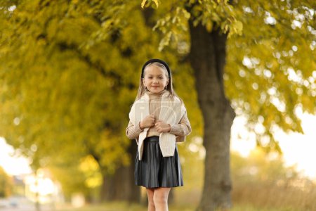 Children's day. Cheerful little child girl 5-6 years old posing and smiling at camera outdoors stand by the tree at autumn park. Stylish tender preschool kid wears beige knit sweater. Happy childhood