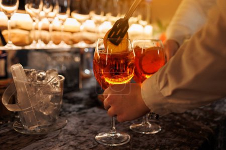 Foto de Cantinero hace dos vasos de cóctel Aperol spritz en el mostrador del bar, añade rodajas de naranja fresca. Bebida típica italiana alcohólica, aperitivo elaborado con vino blanco espumoso Prosecco y cubitos de hielo - Imagen libre de derechos