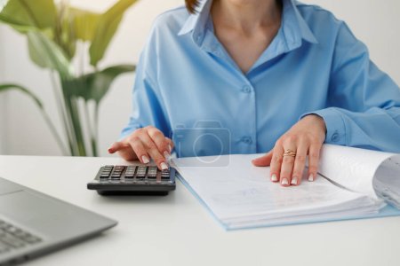 Unrecognisable businesswoman calculates on calculator expenses, fees, analyses financial reports, does accounting, paperwork job, laptop on desk at office. Investment, savings, currency concept.