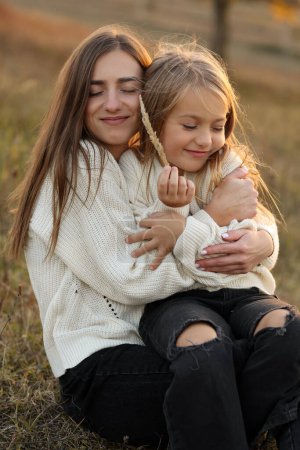 Familientag, Muttertag. Glücklich lächelnde junge Mutter und entzückende Tochter umarmen sich, küssen sich und verbringen Zeit miteinander im Herbst bei Sonnenuntergang. Idyllische Familie vergnügt sich in den Herbstferien draußen