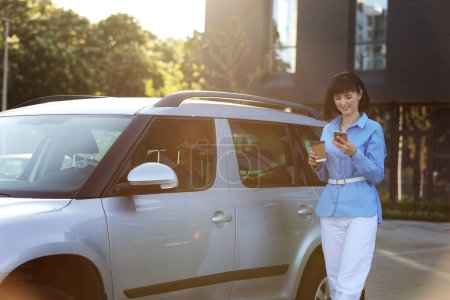 Erfolgreiche Geschäftsfrau in blauem Hemd, weißer Hose mit Pappbecher Kaffee, SMS auf dem Smartphone, während sie am modernen Bürogebäude vorbei zum Auto läuft. Konzept des Selbstmanagements für Produktivität.