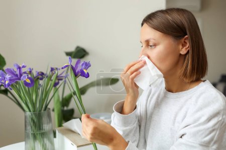 Allergic young woman holds iris flower, covers nose with paper tissue has runny nose, sneezes from flowers pollen at kitchen home by table. Girl with flu, itching or cough, seasonal allergy, rhinitis.