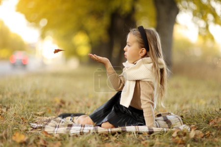 Stylish charming little child caucasian girl 5-6 years has fun outdoors, blows fallen leaves from her hand, sits on plaid on grass outdoors at autumn park. Children's day. Leisure activity on holiday.
