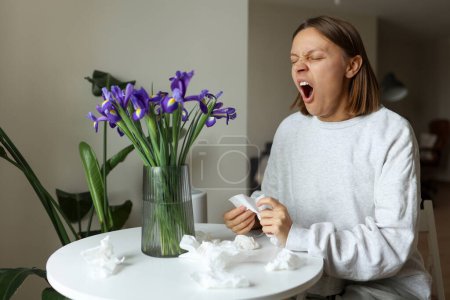 Libre de alergia estacional. Mujer joven huele a flores de iris, disfruta del olfato sin goteo nasal, picazón o tos síntomas estacionales en el hogar acogedor. La chica se sienta junto a la mesa. Tejidos de papel usados en la mesa.