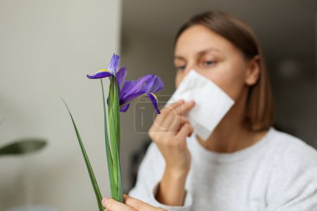 Allergische junge Frau hält Irisblüte in der Hand, deckt Nase mit Papiertaschentuch zu, hat laufende Nase, niest von Blütenpollen zu Hause am Tisch. Mädchen mit Grippe, Juckreiz oder Husten, saisonaler Allergie, Schnupfen.