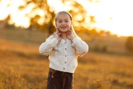 Kindertag. Portrait von entzückenden kleinen kaukasischen Mädchen 5-6 Jahre lächelt und blickt in die Kamera mit gekreuzten Händen ihre Hand im Freien im Herbst Sonnenuntergang Lichter. Freizeitaktivität in den Herbstferien
