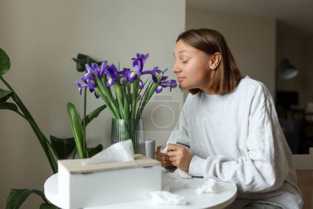 Libre de alergia estacional. Mujer joven huele a flores de iris, disfruta del olfato sin goteo nasal, picazón o tos síntomas estacionales en el hogar acogedor. La chica se sienta junto a la mesa. Tejidos de papel usados en la mesa.