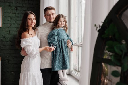 Family day. Happy young mother and father with love embrace little child daughter by window at home, stylish parents in festive clothes hold hands small kid relax rest in living room. Children's day