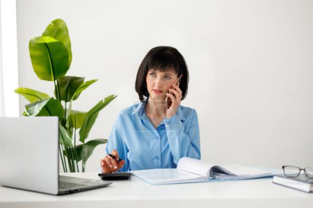 Confident businesswoman communicating with customer by smartphone and counting with calculator at modern office. Laptop and financial documents on work desk. Accountant, auditor, entrepreneur