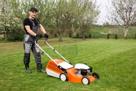 Professional gardener in protective apparel is mowing green grass lawn using modern gasoline cordless lawnmower at backyard. Seasonal landscaping design work. Blooming trees on background.