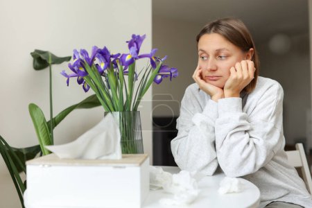 Seasonal allergy. Unhealthy allergic young woman uses paper tissues, suffers from runny nose, sneezes from flowers pollen at kitchen home by table. Girl with flu, itching or cough, allergy rhinitis.