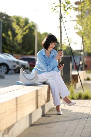 Elegant businesswoman holds paper cup with coffee or tea to go, messaging on smartphone and relaxing free time outside office building. Female adult worker has lunch break in business area on bench.