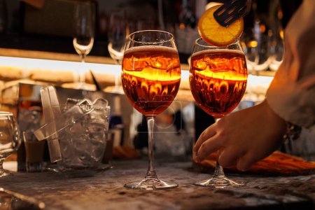 Bartender makes two glasses of cocktail Aperol spritz on bar counter, adds fresh orange slices. Typical alcoholic Italian beverage, aperitif made with Prosecco sparkling white wine and ice cubes