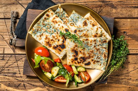 Gozleme flatbread with greens and vegetable salad on garnish. Wooden background. Top view.