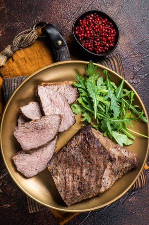 Téléchargez les photos : Rôti Tri Tip ou surlonge steak de bœuf sur une assiette avec roquette. Fond sombre. Vue du dessus. - en image libre de droit