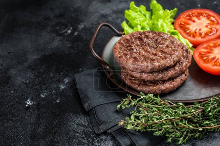 Foto de Tasty grilled burger beef patty with tomato, spices and lettuce in kitchen tray. Black background. Top view. Copy space. - Imagen libre de derechos