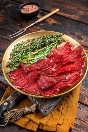 Photo for Basturma, sliced dried beef meat, meat Jerky in steel plate with herbs and spices. Wooden background. Top view. - Royalty Free Image