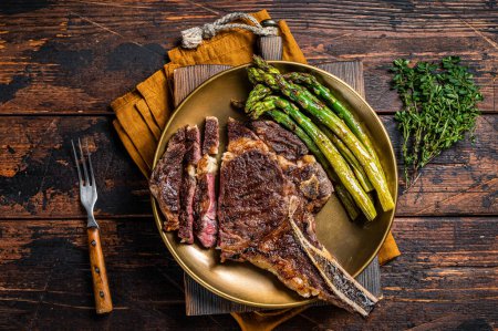 Téléchargez les photos : Sliced Grilled Cowboy or rib eye with bone beef steak, roasted asparagus in a plate. Wooden background. Top view. - en image libre de droit