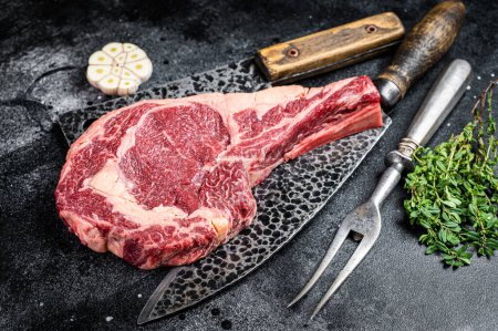 Foto de Dry aged raw tomahawk or cowboy beef steak on a black table with knife, cleaver, spice and herbs. Black background. Top view. - Imagen libre de derechos