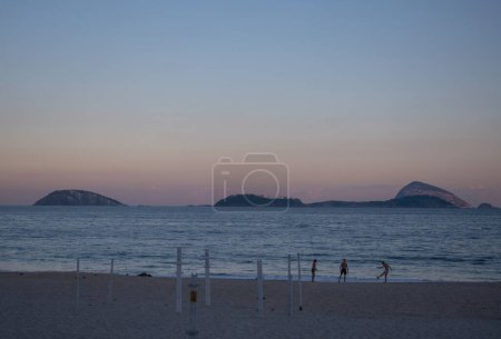 Téléchargez les photos : Rio de Janeiro, Brésil, 07-06-2023 : les gens jouent au coucher du soleil sur la plage d'Ipanema, l'une des plages les plus célèbres au monde, avec le trottoir graphique emblématique le long de l'Avenida Atlantica et les palmiers - en image libre de droit