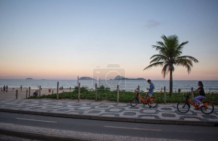 Téléchargez les photos : Rio de Janeiro, Brésil, 06-07-2023 : Vélo au coucher du soleil sur la plage d'Ipanema, l'une des plages les plus célèbres du monde, avec le trottoir graphique emblématique le long de l'Avenida Atlantica (Atlantic Avenue) et des palmiers - en image libre de droit