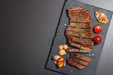 Well done roasting veal steak with sauce, rosemary and grilled garlic. A trendy hard light, dark shadow, black stone concrete background, flat lay, top view