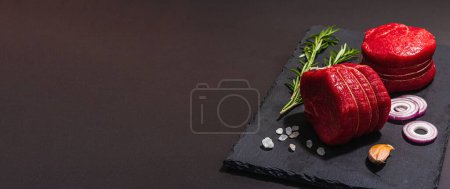 Foto de Ojo crudo de filete redondo con especias y hierbas sobre fondo de hormigón de piedra negra. Una luz dura de moda, sombra oscura, disposición plana, formato de banner - Imagen libre de derechos