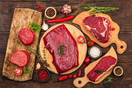 Set of various steaks with traditional spices and herbs. Fresh raw meat cuts includes ribeye, eye round, flank and striploin steaks. Old wooden background, flat lay, top view