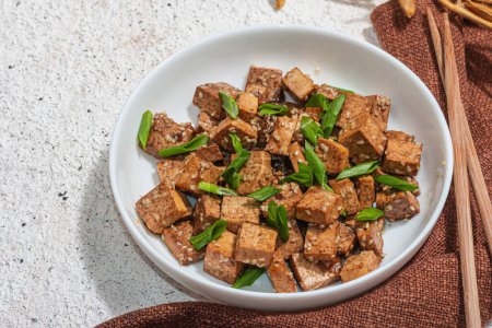 Téléchargez les photos : Tofu tériyaki frit aux oignons verts et graines de sésame. Repas végétalien sain sans viande riche en protéines et en calcium. Lumière dure tendance, ombre sombre, fond de plâtre, gros plan - en image libre de droit