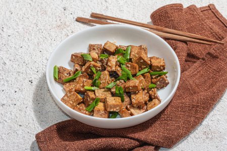 Téléchargez les photos : Tofu tériyaki frit aux oignons verts et graines de sésame. Repas végétalien sain sans viande riche en protéines et en calcium. Lumière dure tendance, ombre sombre, fond de plâtre, gros plan - en image libre de droit