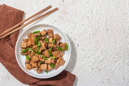 Téléchargez les photos : Tofu tériyaki frit aux oignons verts et graines de sésame. Repas végétalien sain sans viande riche en protéines et en calcium. Lumière dure tendance, ombre sombre, fond plâtre, vue de dessus - en image libre de droit