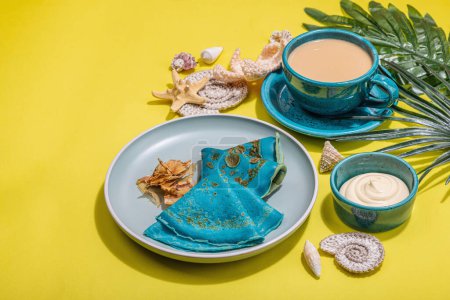 Photo for A cup of coffee and blue thin pancakes in a marine style. Starfish, sea shells, palm leaves. Hard light, dark shadow, bright yellow background, copy space - Royalty Free Image