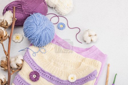 Handmade crocheted baby t-shirt in lilac tones. Stuff and props contains thread, hooks, knitting needles and craft decor. Light stone concrete background, top view
