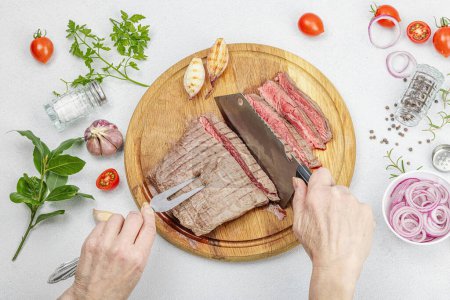 Photo for Woman cuts grilled steak. Female hands with knife and two pronged fork. Fresh herbs, spices, vegetables. Wooden cooking board, picnic or barbecue concept. Light stone concrete background, top view - Royalty Free Image